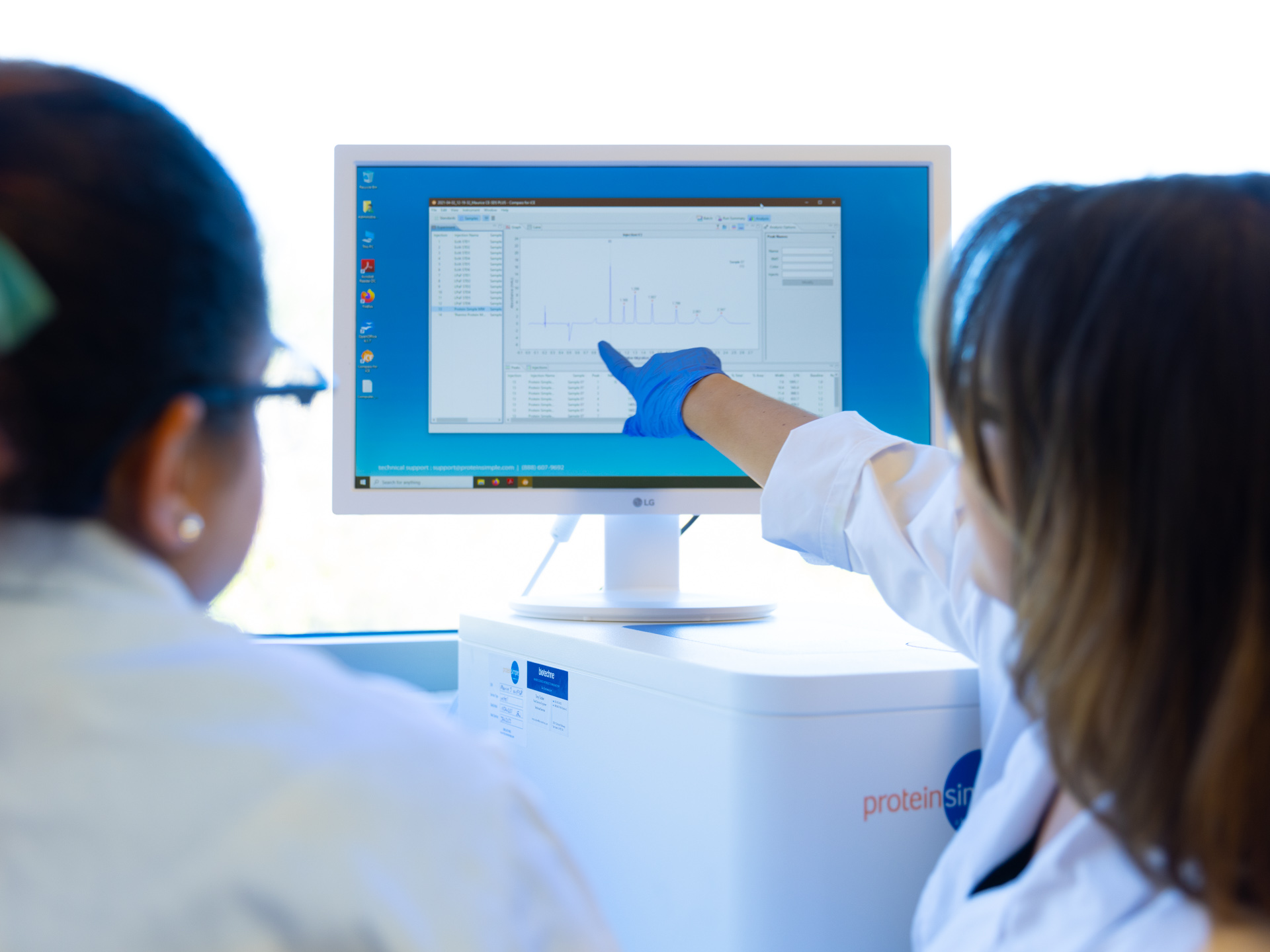 Two female scientists in lab coats review data on a computer about SBIR / STTR grant proposal writing.