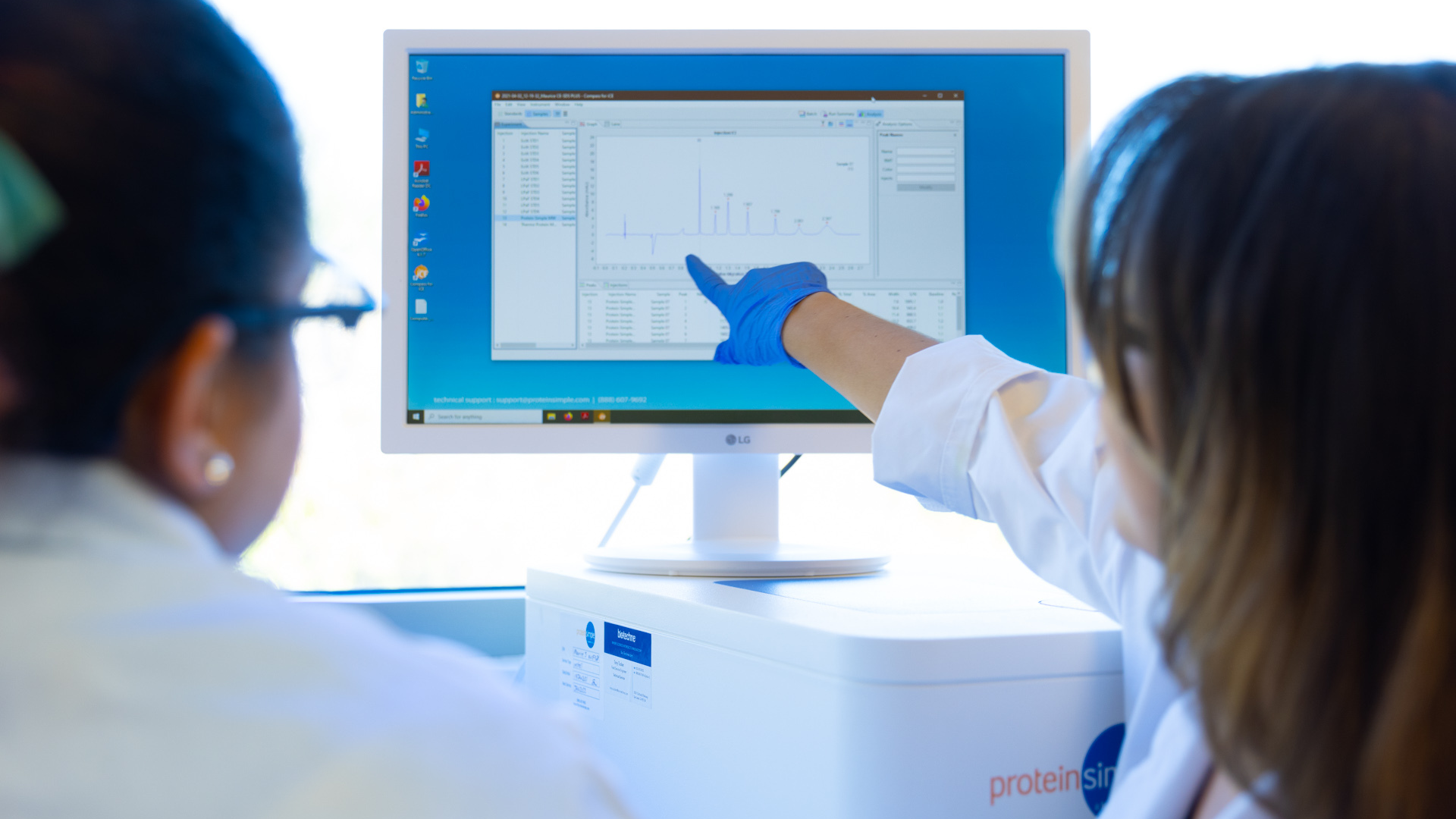 Two scientists in lab coats analyze data on a computer screen. One scientist, wearing blue gloves, points at a graph displaying peaks and numerical data. The screen shows a software interface with analytical results. The setting is a laboratory with a scientific instrument visible below the monitor.